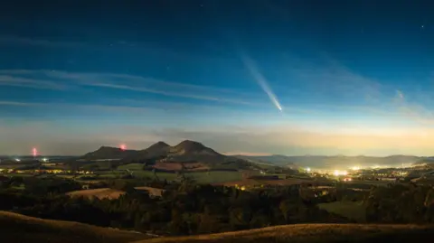 CMuzz/BBC Weather Watchers Comet A3 pictured over hills and a settlement with streetlights.