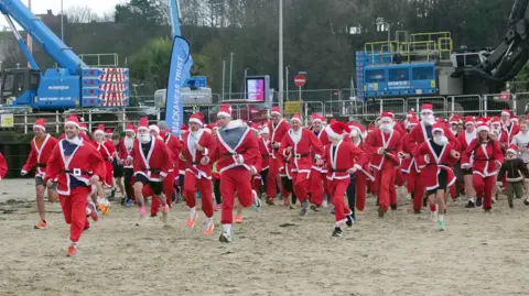 Dozens of runners dressed up like Santas running on Weymouth beach as part of the Chase the Pudding event. It's an overcast day.