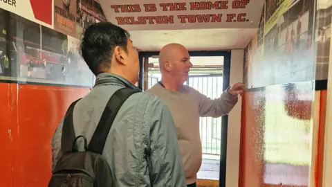 Bilston Town FC A man with dark hair, a pale blue jacket and black backpack in a corridor with orange and white walls leading outside, with a man with a bald head and grey jumper pointing at a wall