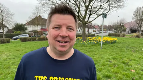 A man smiling to camera wearing a navy t-shirt, he has dark short hair and is standing on a grassy patch by a residential road with daffodils in the background