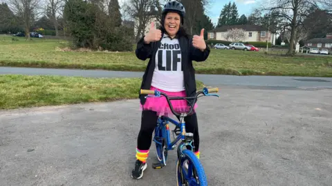 A woman with long dark hair is giving the thumbs-up to the camera as she sits atop a blue bike. She is wearing a cycling helmet, a pink tutu, a "Choose Life" T-shirt, black leggings and brightly-coloured ankle warmers.