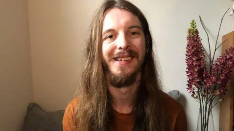 James Moore, who has long brown hair and a brown beard, smiling. There are some flowers in a vase in the background