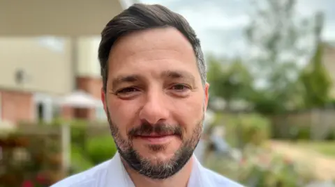 Razvan Stanbeca has short brown hair and a beard and is standing in garden outside of a care home. He is wearing a white shirt.