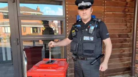 West Mercia Police A police officer wearing a black t-shirt and black vest that says "police" on it, in silver writing on a blue background. There is a walkie talkie on his chest, a body cam, and a baton strapped to his waistband. He is holding a small knife over the entrance of a red knife surrender bin.