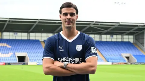 Isaac Fletcher standing with arms folded in dark blue Shrewsbury Town shirt with a stand at Croud Meadow in the background