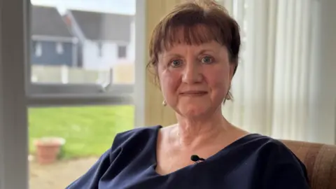 A woman with short ginger hair and a dark blue top in a chair with a large window behind her