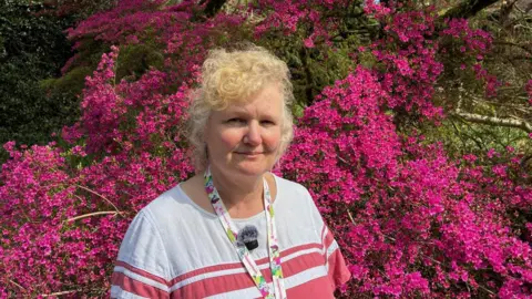 A woman smiles at the camera , she has blonde curly hair . Behind her is a shrub with bright magenta coloured flowers