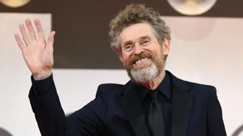 ETTORE FERRARI/EPA/Shutterstock Willem Dafoe wearing an all black suit waving with one hand to the cameras on the red carpet.