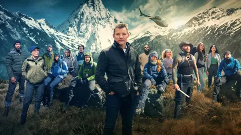 Shine TV A man in a jacket stands centrally with his hands in his pockets. He is surrounded by 14 other people. They are all on a grassy area surrounded by snow-capped mountains with a helicopter hovering in the air.