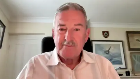 BBC News Andy Wyatt, wearing a white shirt and with grey hair and moustache, sits in a home office looking towards the camera. Pictures of aircraft are visible on the wall behind him.