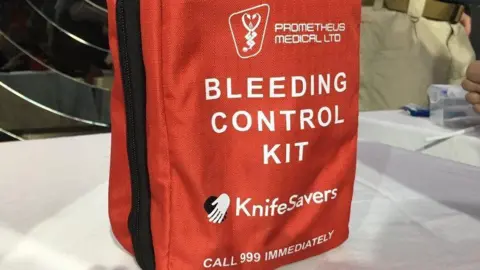 A small red bag with a black zip that reads "Bleeding control kit, Knifesavers, call 999 immediately"