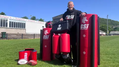 A grey-haired man stands on a sports pitch, surrounded by red and black training equipment