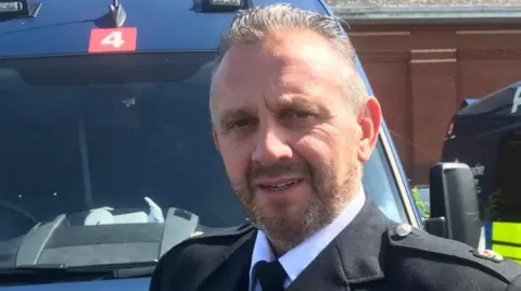 Guernsey Police Damian Kitchen stood in front of a blue vehicle. He is wearing a black jacket with two buttons on each shoulder. He has short hair, a beard and is smiling