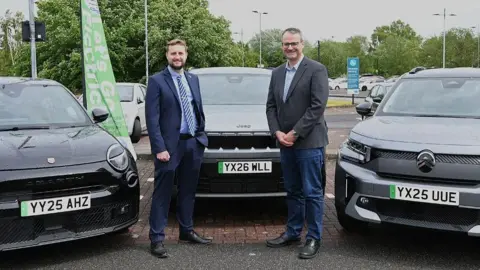 Two men stand in front of a black electric car, with other models to either side, in a car park. The man on the left has brown hair and a beard and is wearing a blue suit and blue and white striped tie. The man on the right has short grey hair and wears glasses, a grey blazer and blue jeans.