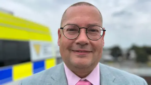 Simon Outen-Coe, wearing a pink shirt and tie under a grey suit and glass, smiles for the camera, with a blurred out police vehicle in the background 