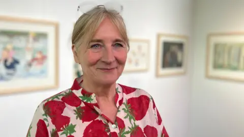 Jon Wright/BBC Dr Amanda Hodgkinson in a shirt with a strawberries design, stands in front of artwork on the walls in a gallery
