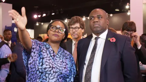 Joyce Liu/BBC David Lammy stands beside Tokini Peterside, a woman wearing a blue dress with a leaf print and black glasses, as she points towards something off camera