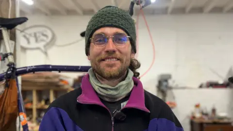 Alan is smiling at the camera. He is standing in a workshop with his tall bike behind him. He is wearing round glasses, a green beanie hat and a colourful jumper. 