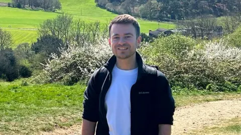 Jordan Waite A man stands in the countryside. He is looking at the camera. He has short dark brown hair and is wearing a white top and a dark jacket with the sleeves rolled up