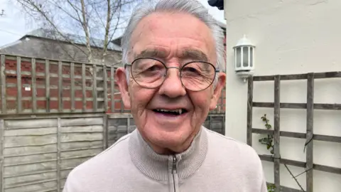 A man wearing a beige jacket with a zip stands in a garden.