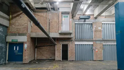 An empty storage warehouse, with brick walls. A blue fire exit door is on the left, with metal shutters on the right. A pipe runs across the width of the room. 