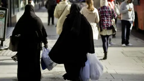 Getty Images Dua wanita yang mengenakan jilbab hitam berjalan menyusuri jalan di London membawa tas belanja plastik.