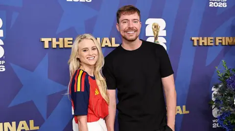 Mr Beast on the right is wearing a plain black t-shirt and black trousers. Thea Booysen is wearing a red, navy blue and yellow football shirt. She has long blond hair and blue eyes. 