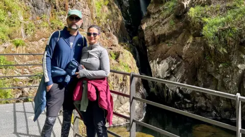 Geet Athwal A man and a women are standing on a metal bridge with rocks and waterfalls behind them. The woman has dark hair and sunglasses, she is wearing a grey top and black trousers with a pink jacket and green hoodie tied around her waist. The man has blue cap on and has grey and black facial hair. He is wearing a blue zip jacket and has a blue coat tied around his waist