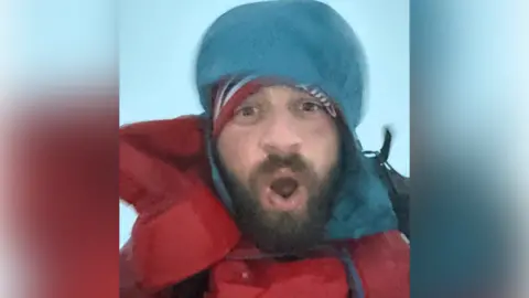 Jake Evans A man dressed in hiking gear including large red coat over a blue hooded-top. He is taking a selfie while his mouth is wide open. Foggy conditions can be seen in the background.