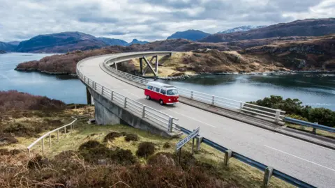 A red and white campervan crosses the Kylesku Bridge. The bridge is set in a Highland landscape of hills and lochs.