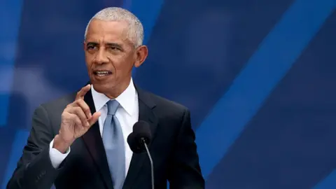 Barak Obama in a dark suit, white shirt and grey tie. He is standing in front of microphones, against a dark blue background. 