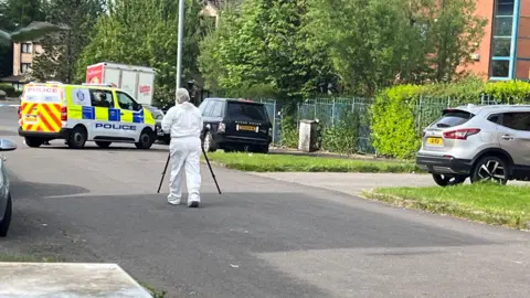 a forensics officer in white overalls on a street with a police van and parked cars
