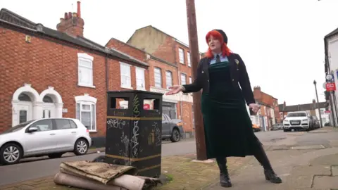 Ant Saddington/BBC Khandie stands next to a street bin, which has discarded carpet next to it, holding one hand towards it. She has bright red hair and wears a hat, a velvet green dress and black knee boots. 