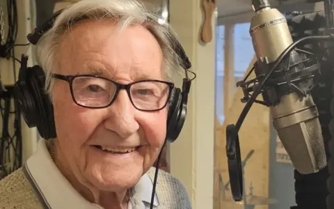 Mr Rayner looking at the camera and smiling. He has brown eyes and white hair. He is wearing headphones and is sitting in front of a microphone.