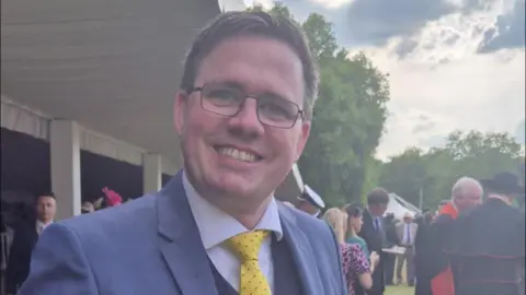 Ryan Davies/Facebook A smiling Ryan Davies has short hair and is wearing glasses. He has a light blue suit jacket, blue shirt and yellow spotted tie. He is pictured at an outdoor event with other attendees in the background. 
