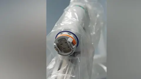 Royal Cornwall Hospitals NHS Trust A clear wrapping which looks like plastic surrounds a thin robotic arm with a depression at the end of it and what appear to be controls.