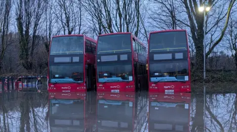 Damory Three red buses lined up in a flooded car park on a dark day. There are some cars behind the busses.