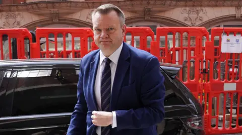 PA Media Nick Thomas-Symonds has short grey hair, he is wearing a navy suit and tie, with white shirt, and is pictured looking serious with a black car and red plastic railings behind him