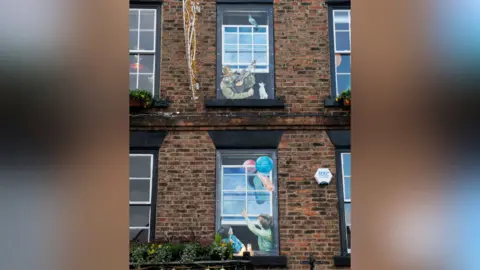 North Yorkshire Council A top-floor window painting depicts a man with a cat and a fiddle. In a window painting directly below, two people point up towards the man.