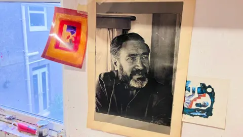 A black and white head shot of Ronald who has dark hair which is brushed off his face and a full beard. He is wearing a black short. The photo is on the wall of her studio and is next to a window covered in open oil paint tubes. 