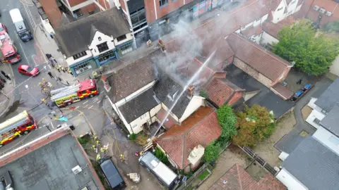 Jayden Stracey An aerial photograph above the Cat and Fiddle pub where a fire has taken place. A hose is being used to put out the fire and smoke is coming from the roof of the building.
