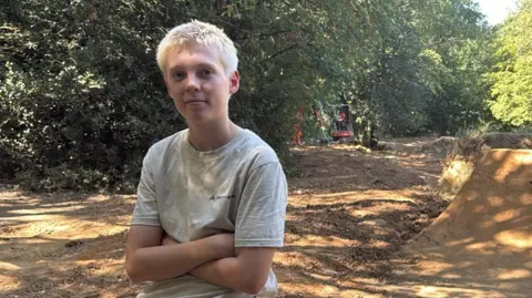 DUNCAN HILL/BBC Walter is sitting on a raised dirt bank with arms crossed. He is wearing a light-coloured T-Shirt and dark shorts.