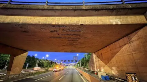 A concrete bridge over the M42 motorway. It has obvious wear and tear and marks on it. The motorway is closed with only highway vehicles parked up with their lights on. 
