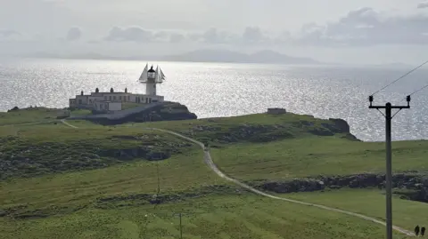 Aldo San Giorgi A remote coastal lighthouse on a grassy headland, overlooking a bright, sunlit sea, viewed from an elevated position.