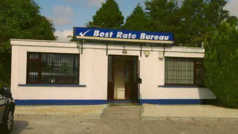 Trainor's former bureau de change business premises on the Dublin road in Newry.  The small one-story building has a flat roof and two rooms on either side of the front door.   A large blue and white business sign above the building says: "Best Rate Bureau". There are security bars on the large, brown- framed windows. 