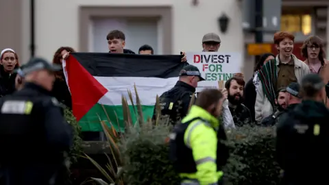 PA Media A group of pro-Palestinian protesters standing behind a wall and fence, holding Palestinian flags and banners. Police officers are seen patrolling and clashing with protesters.