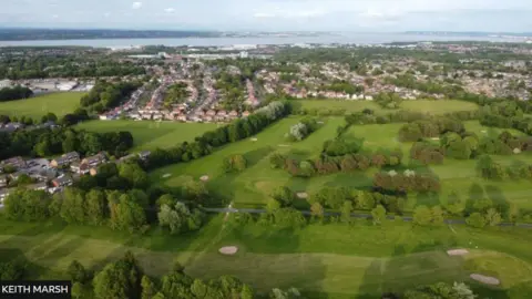 Keith Marsh An aerial shot of the golf course, with numerous trees and holes seen and some housing to the left and behind the course, and the River Mersey in the distance