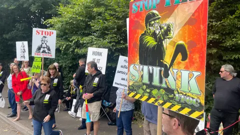 About 15 people protesting against Walleys Quarry. Many are holding placards saying "Stop The Stink" as they stand on the pavement, with a hedge behind them