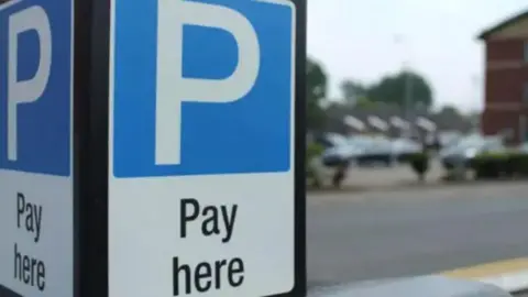 A blue and white parking sign with a white letter P written largely at top with 'Pay here' underneath. There are out of focus cars in the background to what looks like a car park