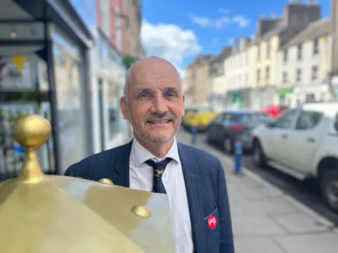 A bald-headed man, wearing a white shirt, dark tie and dark jacket stands behind the golden top of a post box